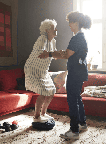 A female physiotherapist holds the arms of an older patient while she balances on a rubber disc. 